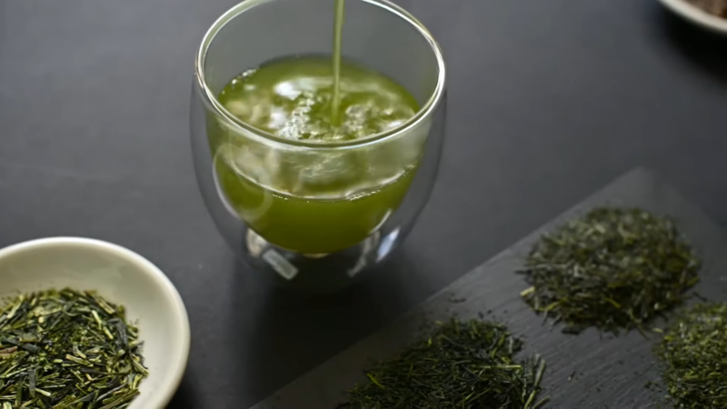 A Glass of Green Tea Is Being Poured, Surrounded by Loose Green Tea Leaves on A Dark Surface