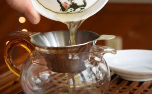 Hot tea is poured through a fine-mesh strainer into a clear glass pitcher