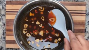 A Hand Stirring a Pan of Dark Sauce with Fried Garlic Slices and Red Chili Pieces Using a White Spoon