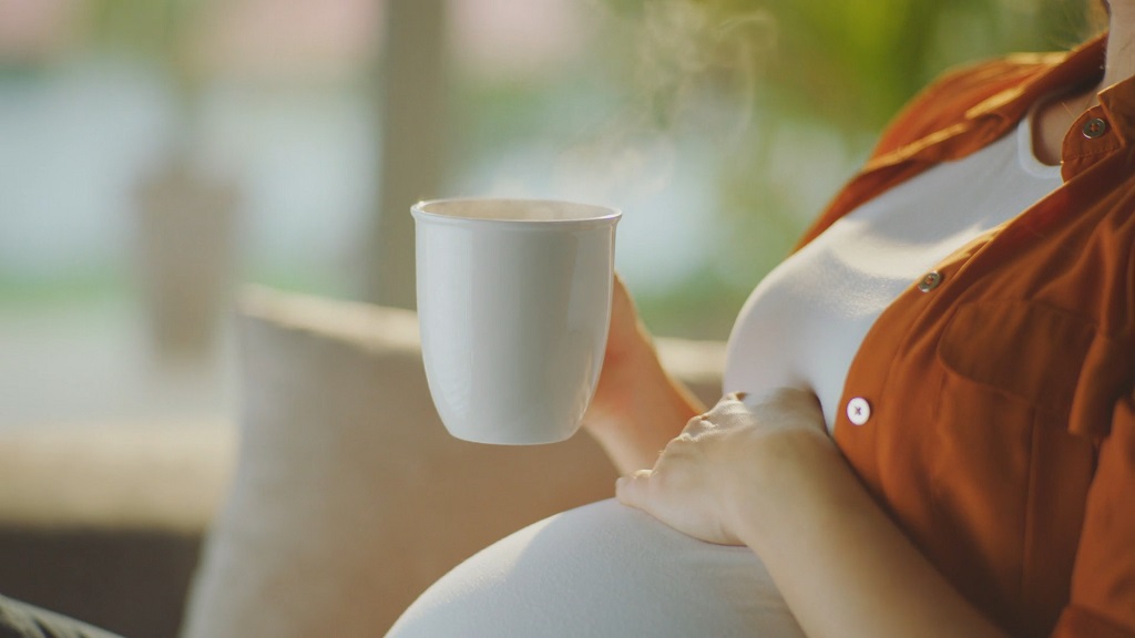 Pregnant woman holding a steaming white mug of tea while resting her hand on her belly