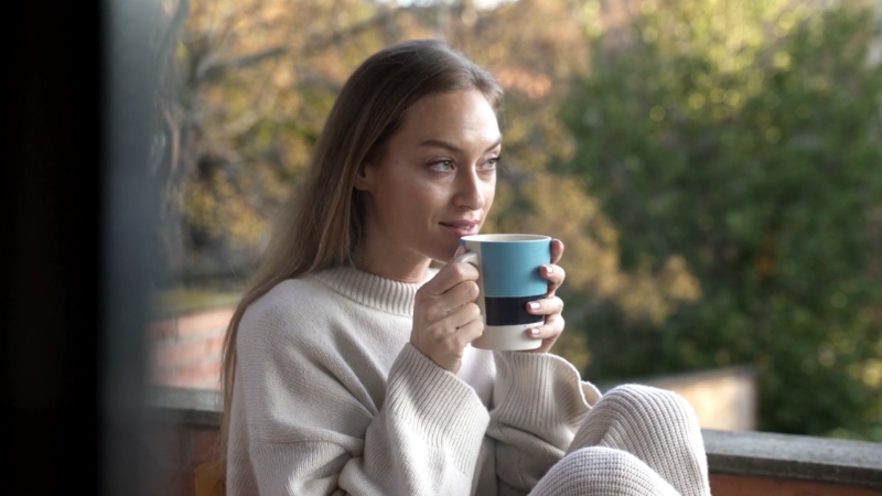 A woman in a cozy sweater sits on a balcony holding a cup of coffee and looking out at the trees